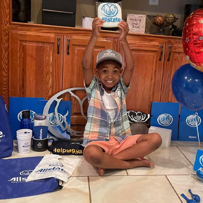 Young boy sitting down surrounded by Allstate swag gifts and holding up an Allstate branding gift with both hand. 